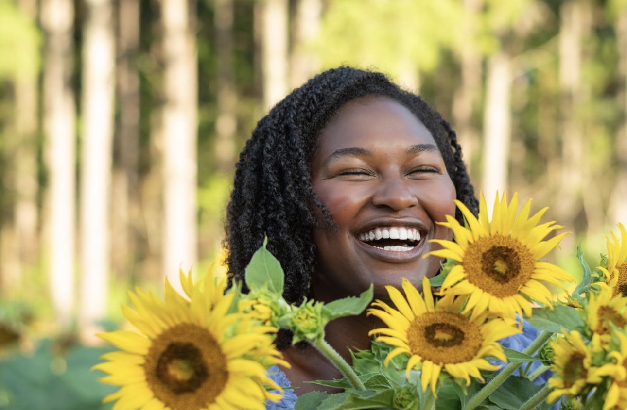 Precious Neely laughing joyfully among sunflowers in a soft lavender-blue dress
