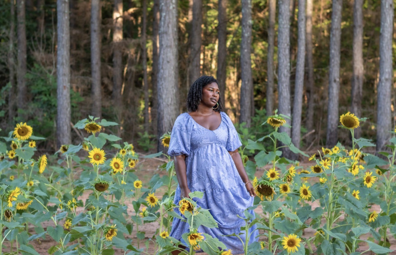 Precious Neely standing peacefully in a golden sunflower field wearing a soft lavender-blue dress